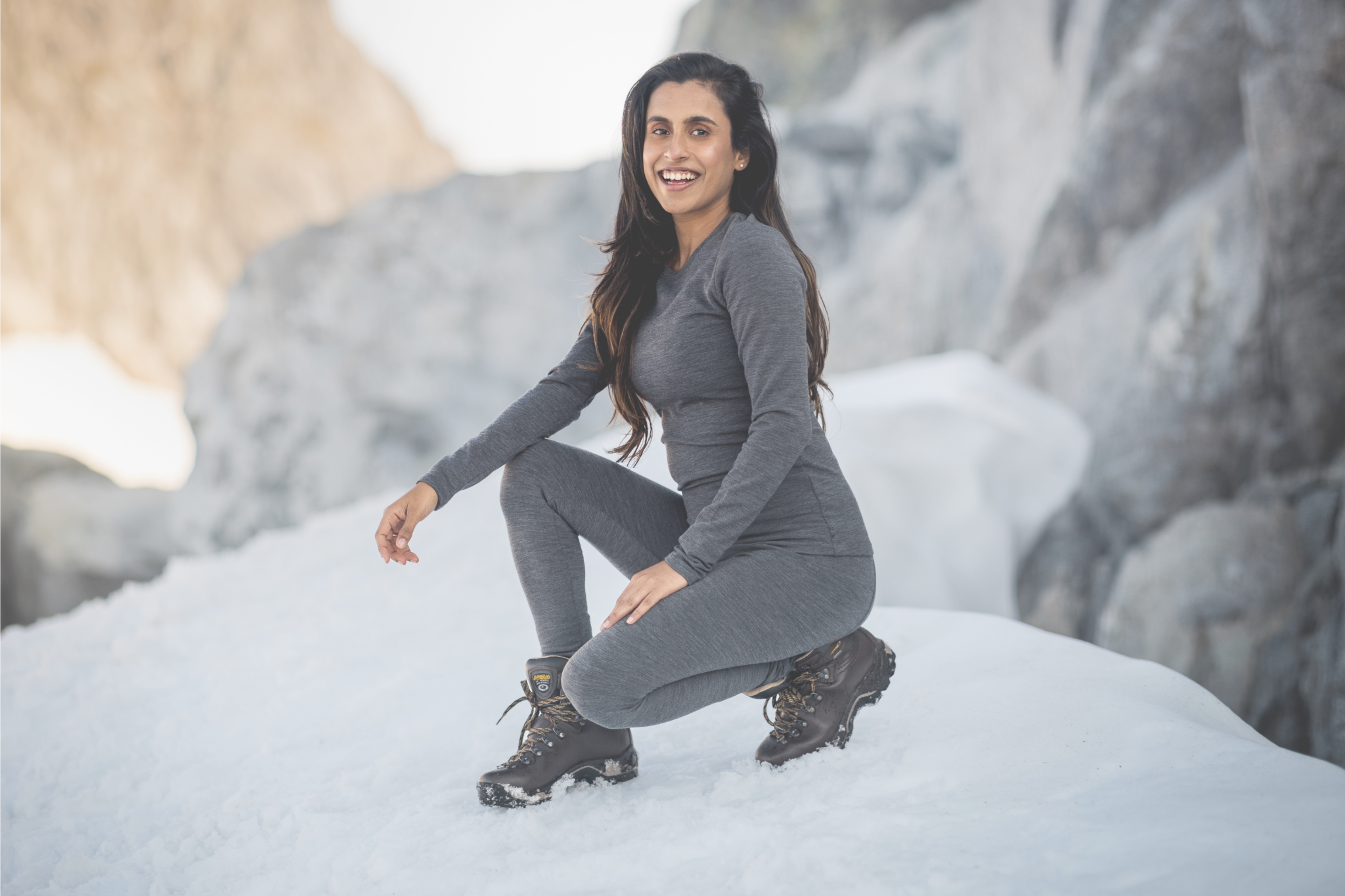 girl squatting down on snow wearing grey top and bottom.  snow covered rocks in background 