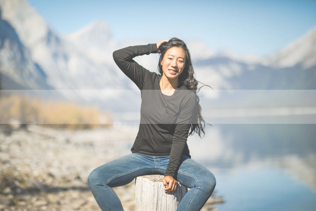Woman sitting on tree stump wearing black long sleeve shirt and jeans, water and mountains in background, looks bright 