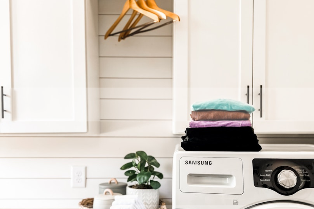 laundry room with clothes folded and set on top of samsung white washer.  hangers for clothes empty hanging above on white cabinets.  fake green plant in corner for decoration.  