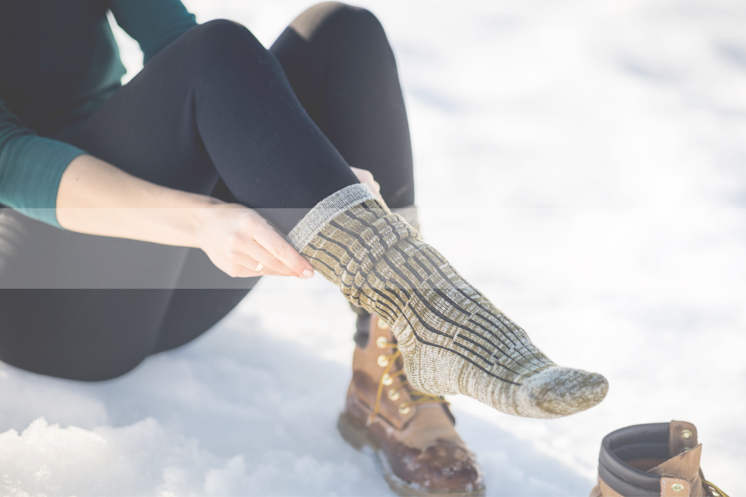 girl sitting in snow, pulling on green and black socks.  one boot on and one off.  