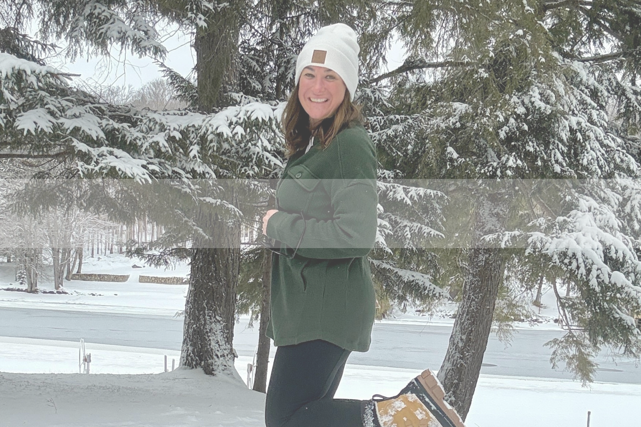 Woman standing in front of 2 pine trees covered in snow, wearing green jacket, white hat and black leggings, posing happy in the snow 