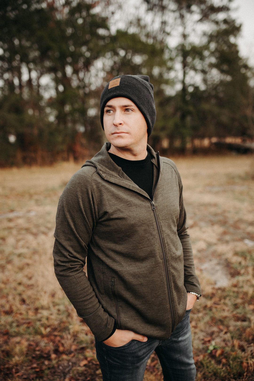 A man wearing a dark beanie and a green zip-up jacket stands outdoors with his hands in his pockets, gazing into the distance. He is surrounded by a natural setting with trees and grass in the background.