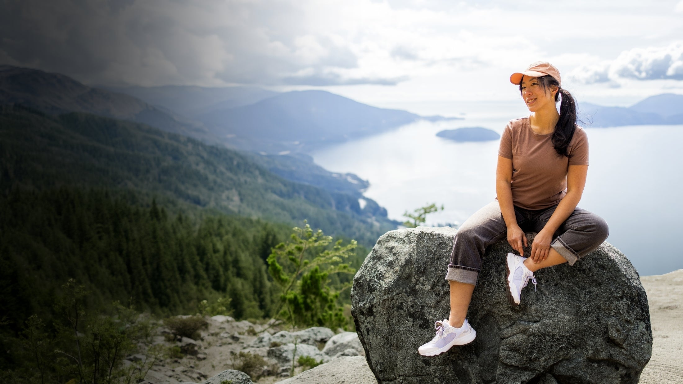 A woman in a cap and casual clothes sits on a large rock, smiling, with a scenic view of forested mountains and a lake in the background under a cloudy sky.