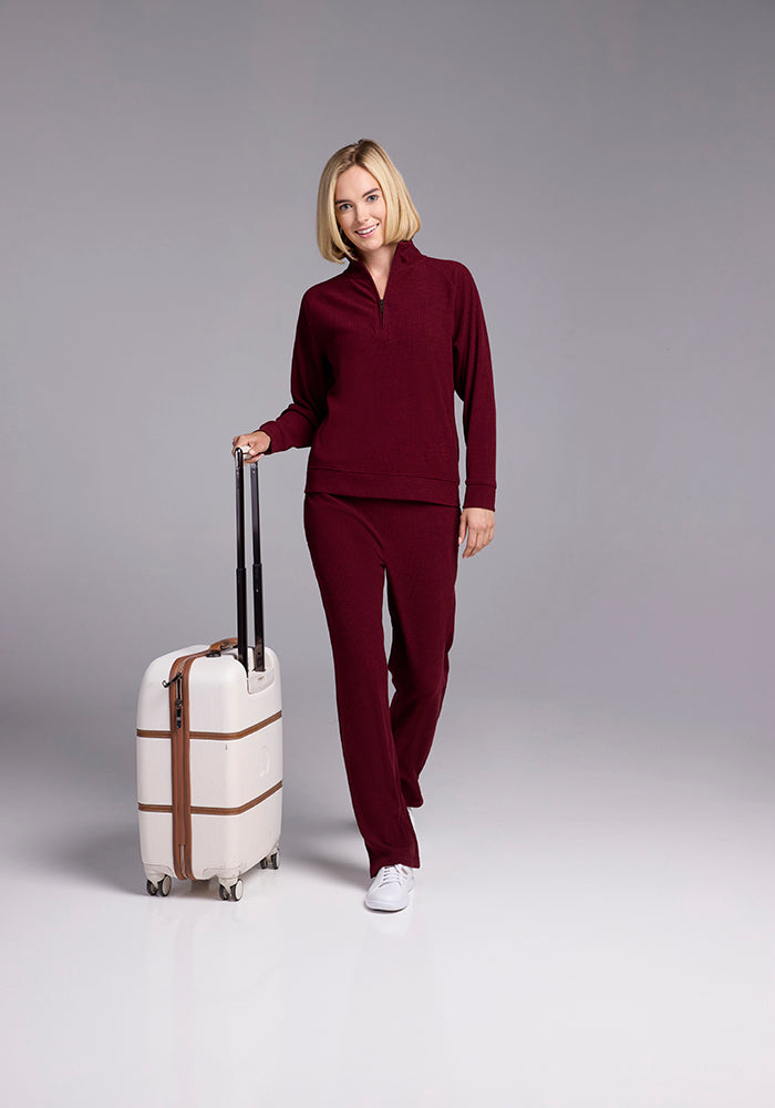 A woman with a blond bob haircut, wearing a burgundy lounge set and white sneakers, stands smiling and holding the handle of a beige rolling suitcase with brown straps against a plain background.