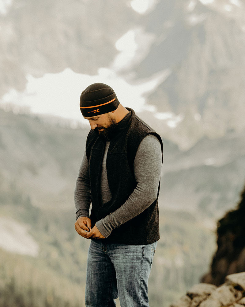A man standing outdoors in a mountainous area, zipping up his black vest. He is wearing a grey long-sleeve shirt, blue jeans, and a black and orange beanie. Snowy peaks and rugged terrain form the backdrop.
