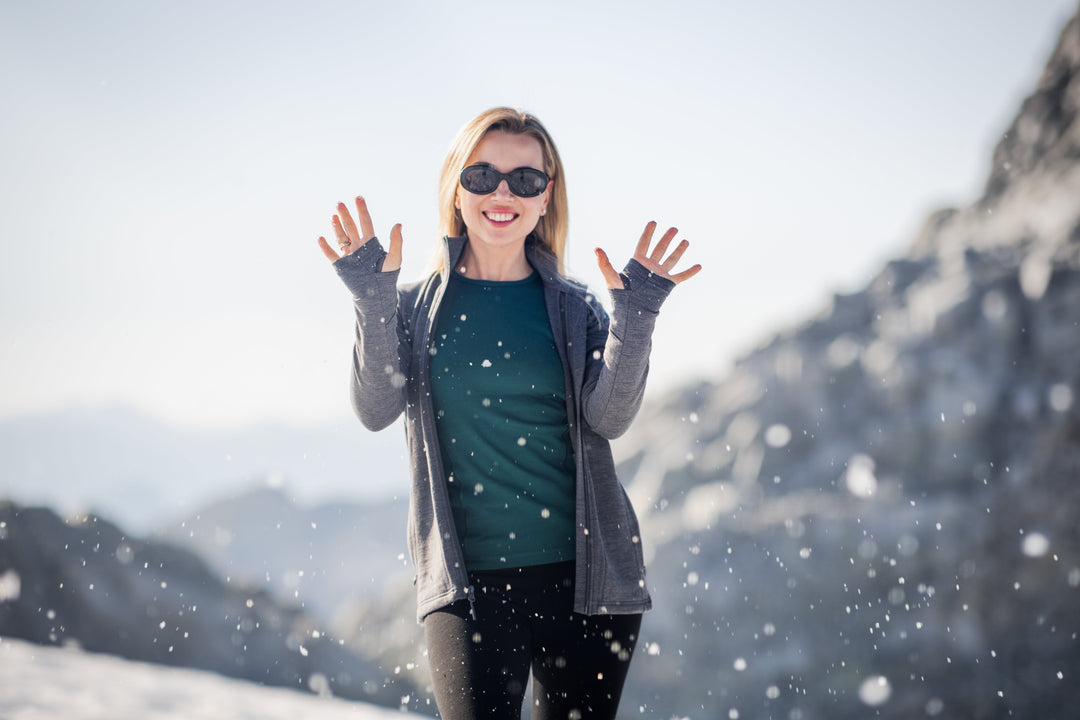 A woman wearing sunglasses and a gray jacket smiles and raises her hands while standing in a snowy landscape with mountains in the background and snow falling around her.