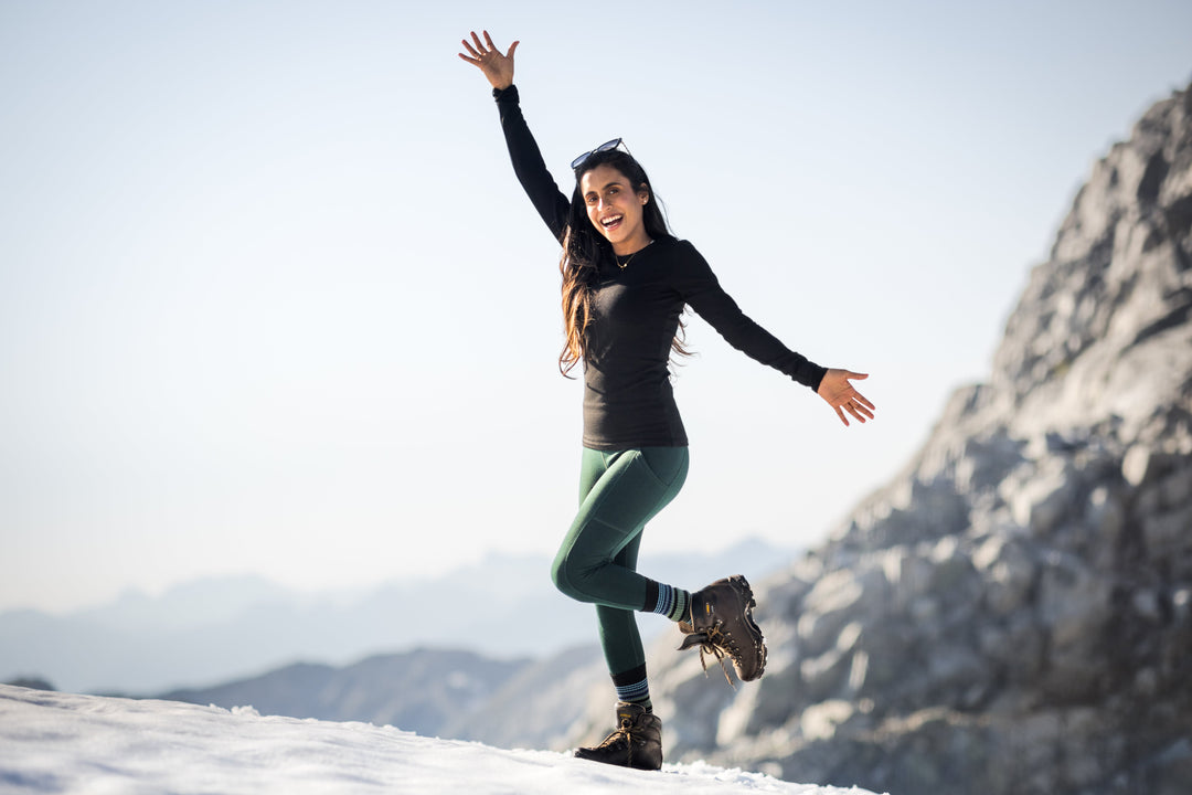 A woman in hiking boots and outdoor clothing stands on snowy ground, smiling with arms raised and one leg lifted. Rocky mountains and a clear sky are visible in the background.
