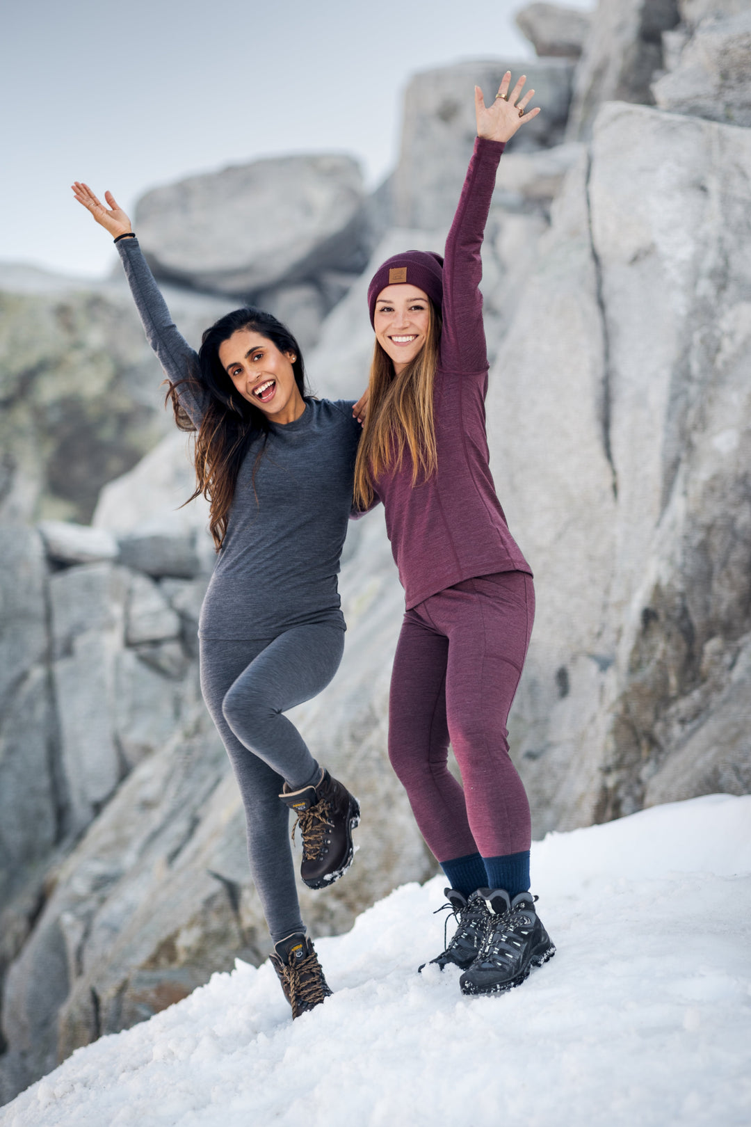 Two women in warm clothing and hiking boots stand on snow with large rocks behind them, smiling and raising their arms joyfully, celebrating outdoors in winter.