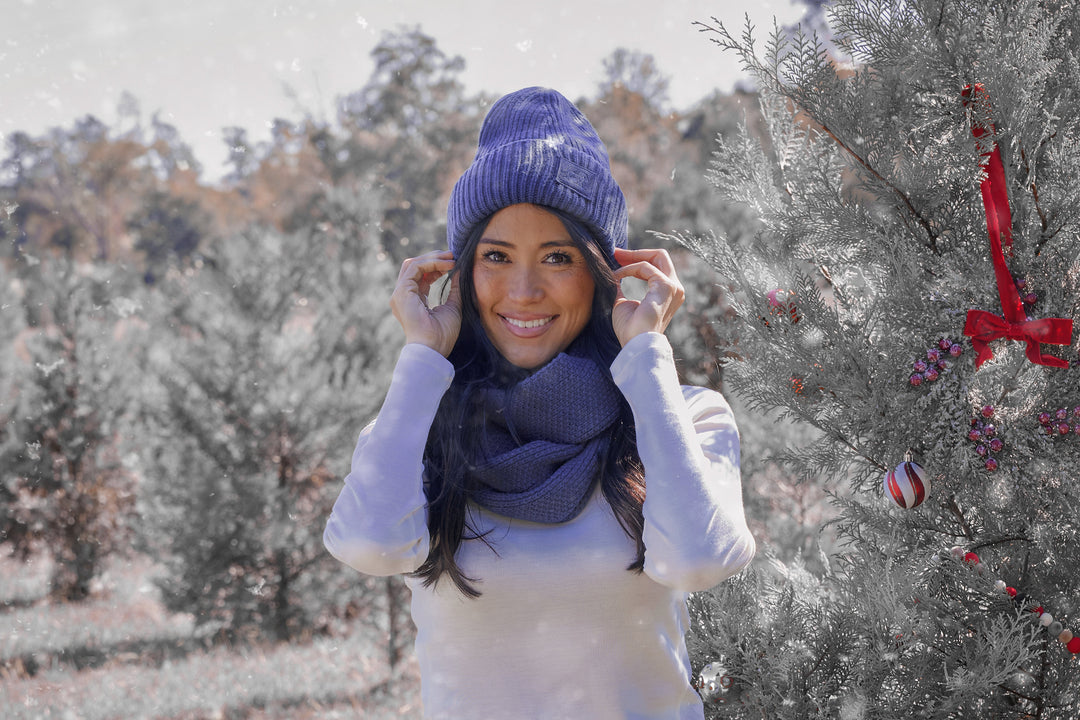 A woman in a blue knit hat and scarf smiles while standing outdoors by a decorated Christmas tree, with snowy trees and a festive winter landscape in the background.
