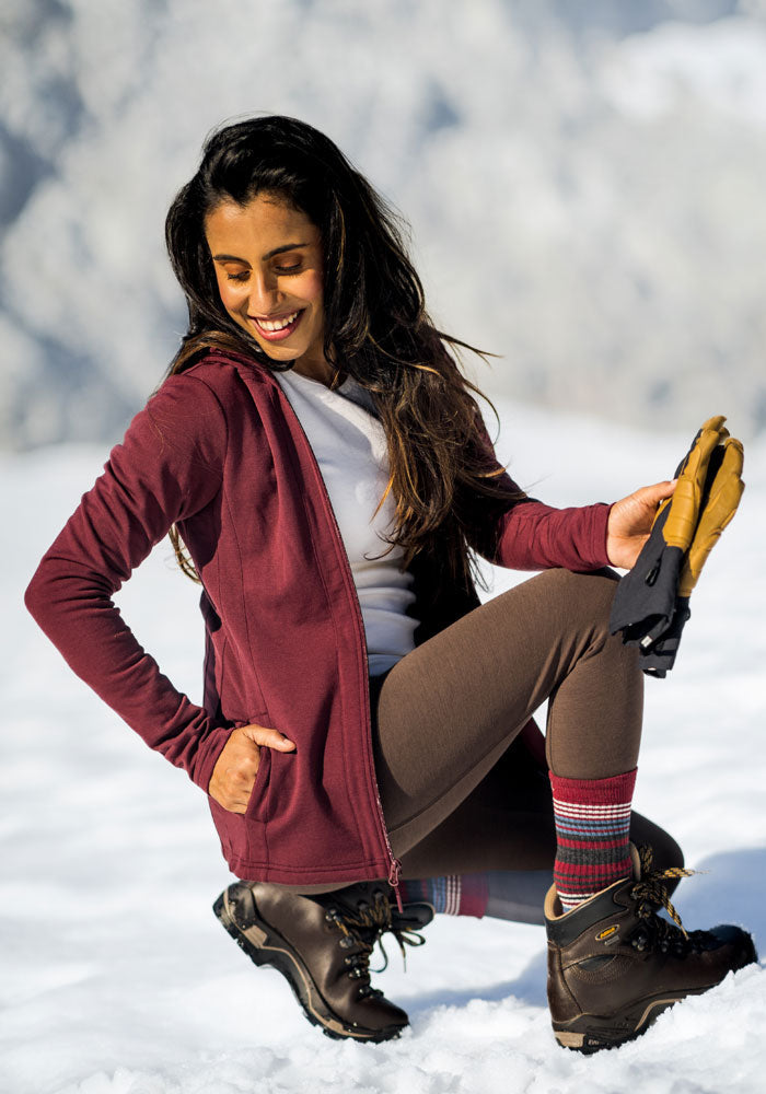 A woman in a maroon jacket and brown hiking boots kneels in the snow, smiling while holding gloves, with a snowy landscape blurred in the background.
