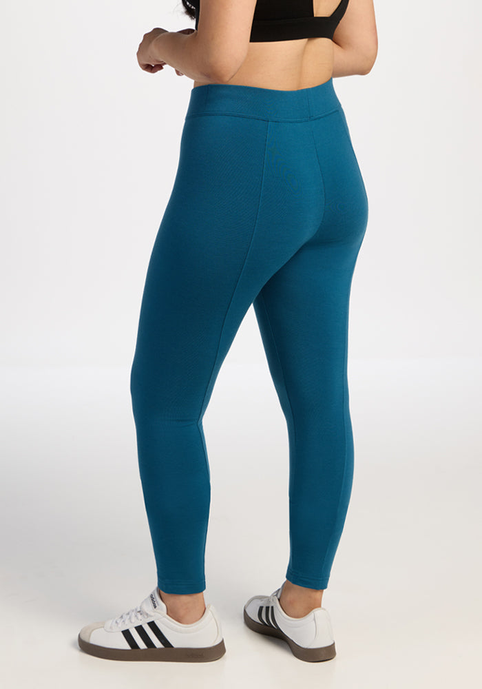 Woman in Woolx Stella Leggings - Blue Pine, black sports bra, and white Adidas sneakers stands facing away on white.