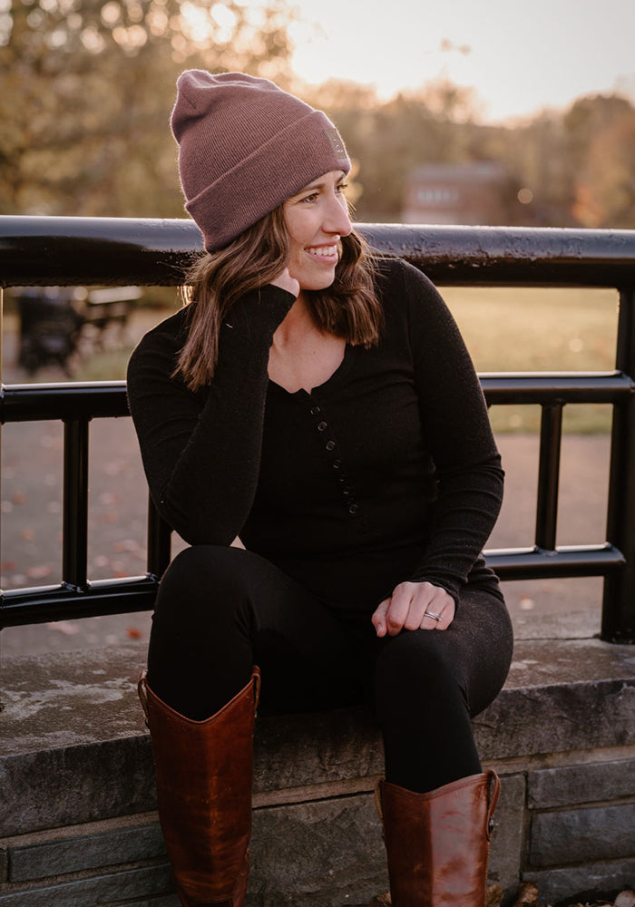 Woman in a Woolx Baylor Beanie - Sable Heather sits on a stone ledge, smiling and looking to the side.