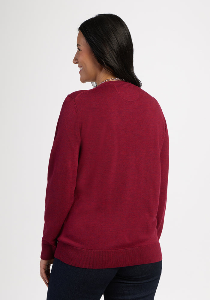 Person wears a Woolx Francesca Sweater in Beet Red, smiling, with dark hair and pants against a white background.