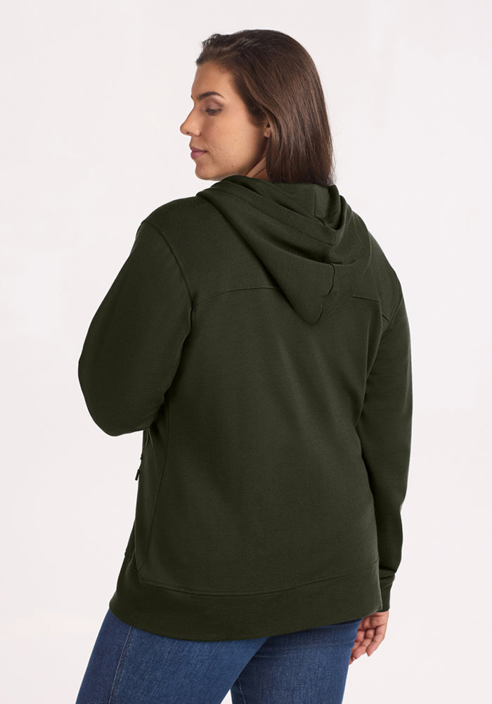 Woman in Woolx Darby Zip Up Hoodie in Duffel Bag and blue jeans, looking over her shoulder against a plain background.