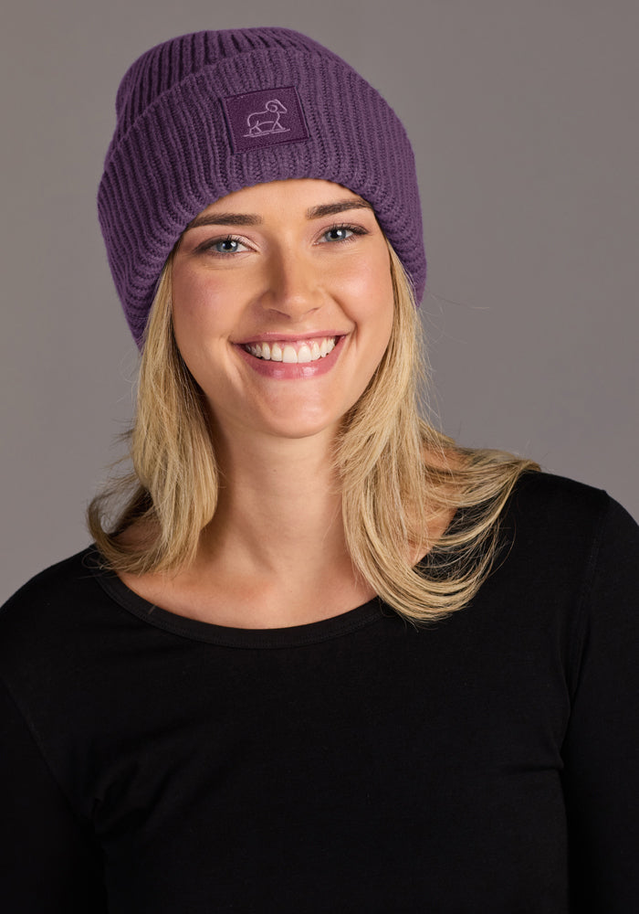 Woman in Woolx Ria Chunky Beanie - Purple Iris and black top, posing against a gray background.