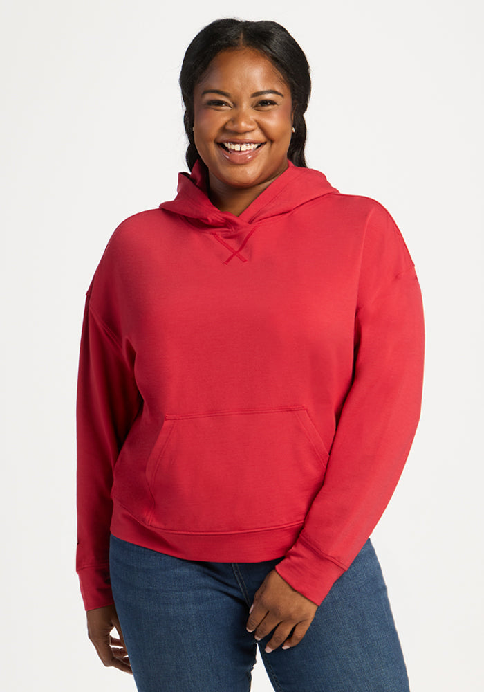 Woman smiling in a Woolx Lizzy Hoodie - Maraschino Cherry and blue jeans, standing against a white background. 