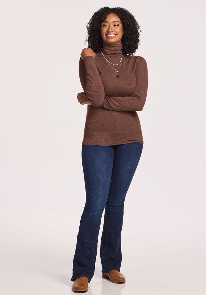 Woman in Woolx Sage Ribbed Turtleneck - Sable Heather, blue jeans, and brown shoes smiles with arms crossed.