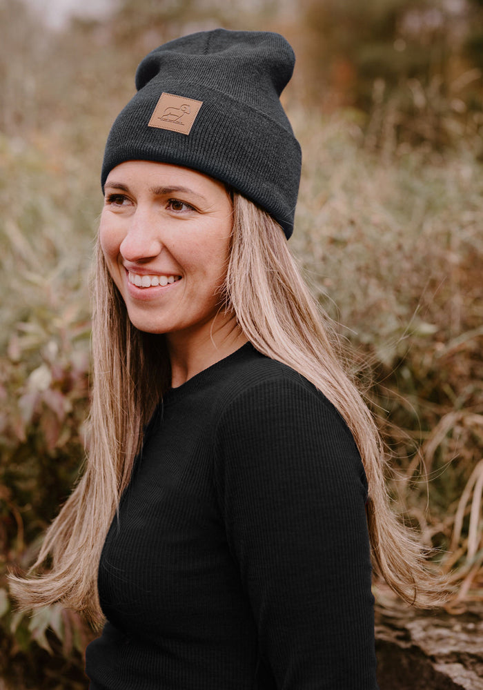Smiling woman wears the Woolx Baylor Beanie in Cinder Melange, standing outdoors with greenery behind her.
