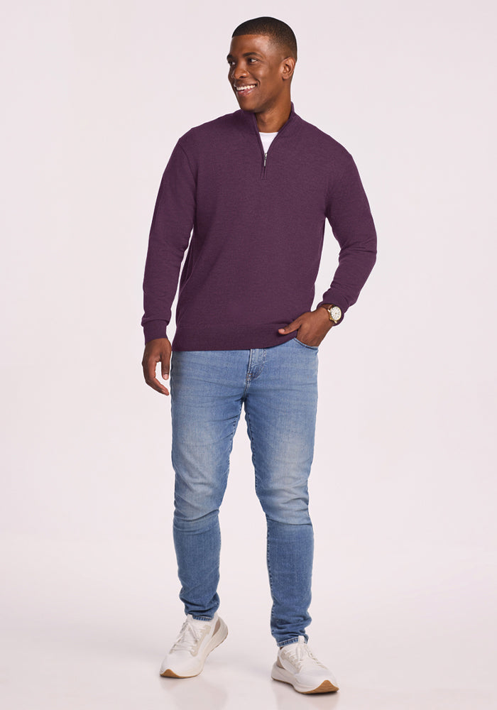 Smiling man in Woolx Enzo Quarter Zip - Purple Iris, blue jeans, and white sneakers stands against a plain background.