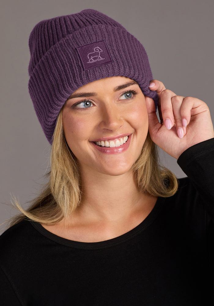 Woman in a Woolx Ria Chunky Beanie—Purple Iris—and black shirt, touching her hat against a gray background.