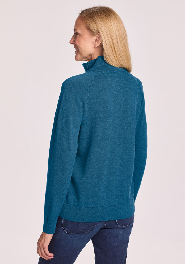 Smiling woman with long blonde hair wears Woolx Collins Half Zip Sweater - Blue Pine and dark jeans, facing slightly away.