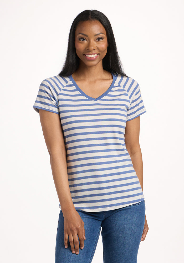A woman with long dark hair smiles in a Woolx Mia V Neck - Blue Sand Stripe short-sleeved, moisture-wicking shirt and blue jeans, standing against a plain white background. 