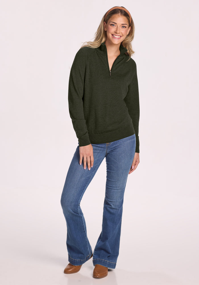 Smiling woman wears a Woolx Collins Half Zip Sweater in Duffel Bag with blue jeans and brown shoes on white background.
