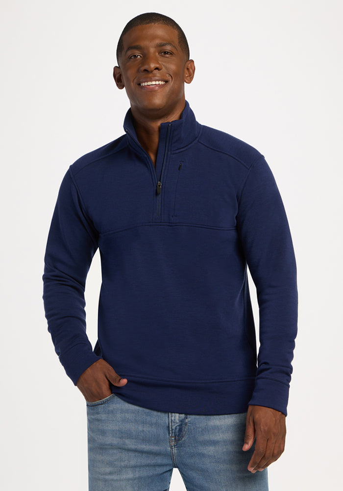 Smiling man in Woolx Mason 1/4 Zip - Ocean Cavern and jeans stands with one hand in his pocket against a plain background. 