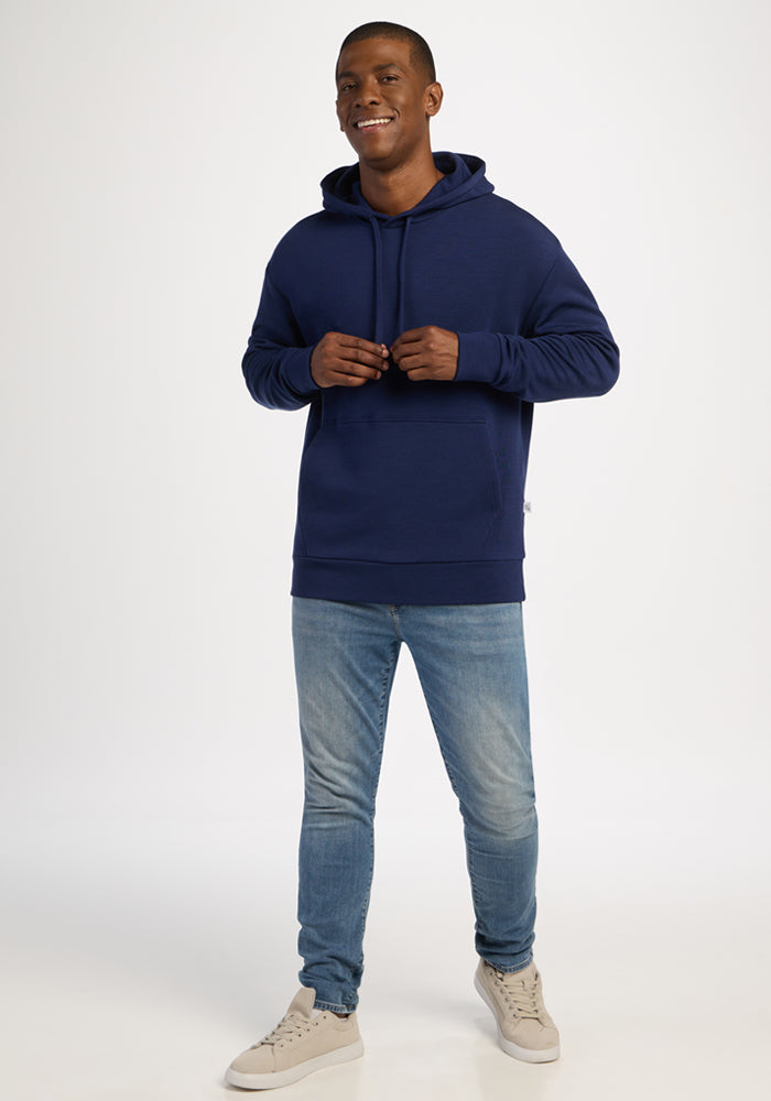 Smiling man in Woolx Watson Pullover - Ocean Cavern, blue jeans, beige sneakers stands against a white background.