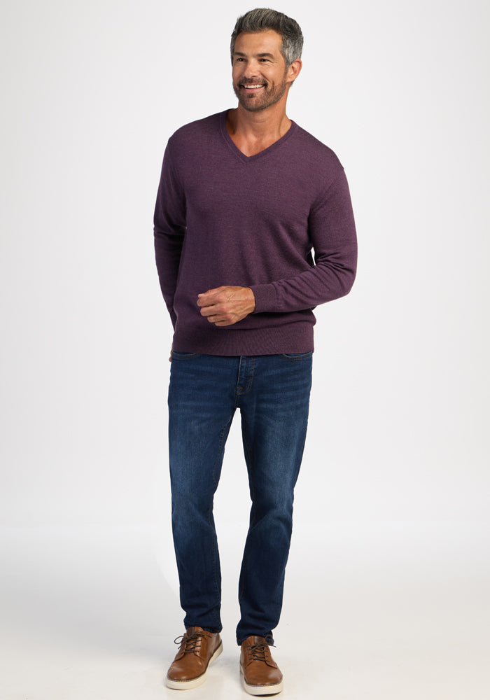 Man wears Woolx Vincenzo Sweater in Purple Iris, blue jeans, and brown shoes against a plain white background.