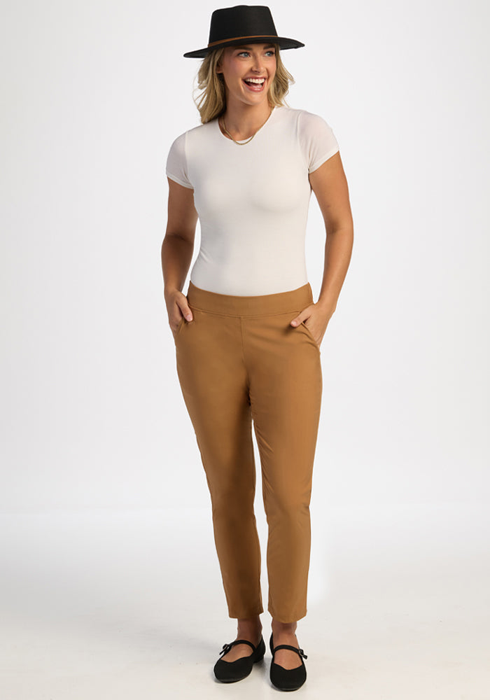 Woman in Woolx Nicole Tall Ankle Pant - Saddleback, white tee, black hat, smiles with hands in pockets.