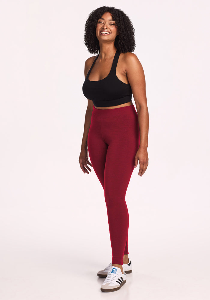 Woman smiles in Woolx Stella Leggings - Pinot Noir, black sports bra, and white sneakers against a plain background.