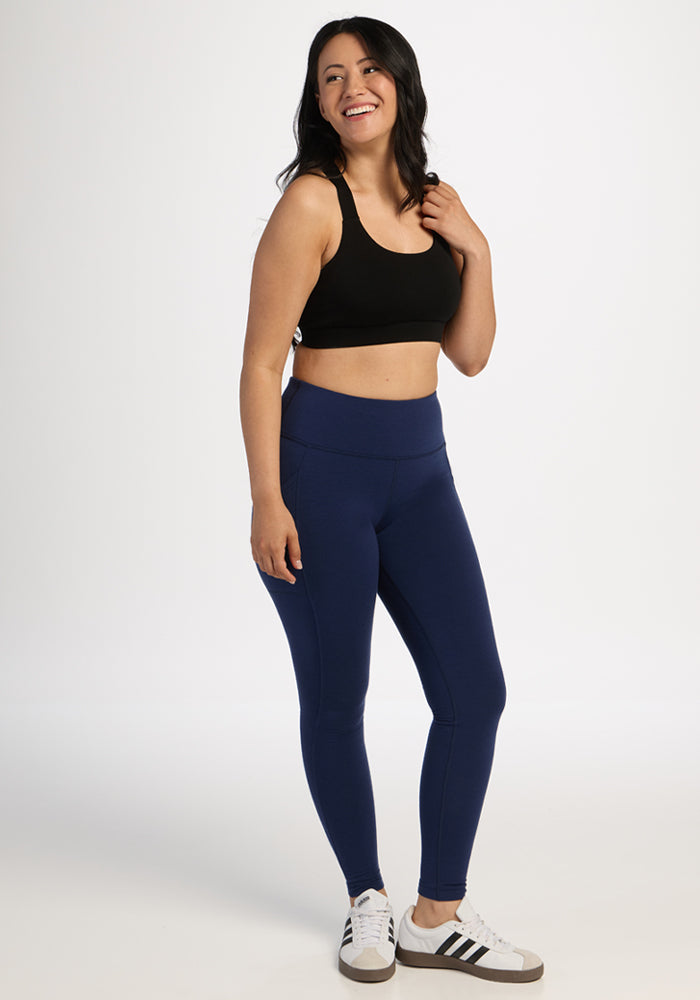 Woman smiling in Woolx Piper Pocket Petite - Ocean Cavern leggings, black sports bra, and white sneakers on a white backdrop.