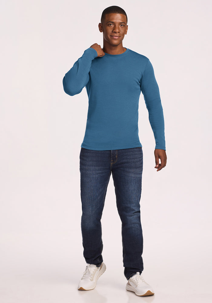 Man in a blue Woolx Easton Crew Neck Shirt, dark jeans, and white sneakers smiles against a plain background.