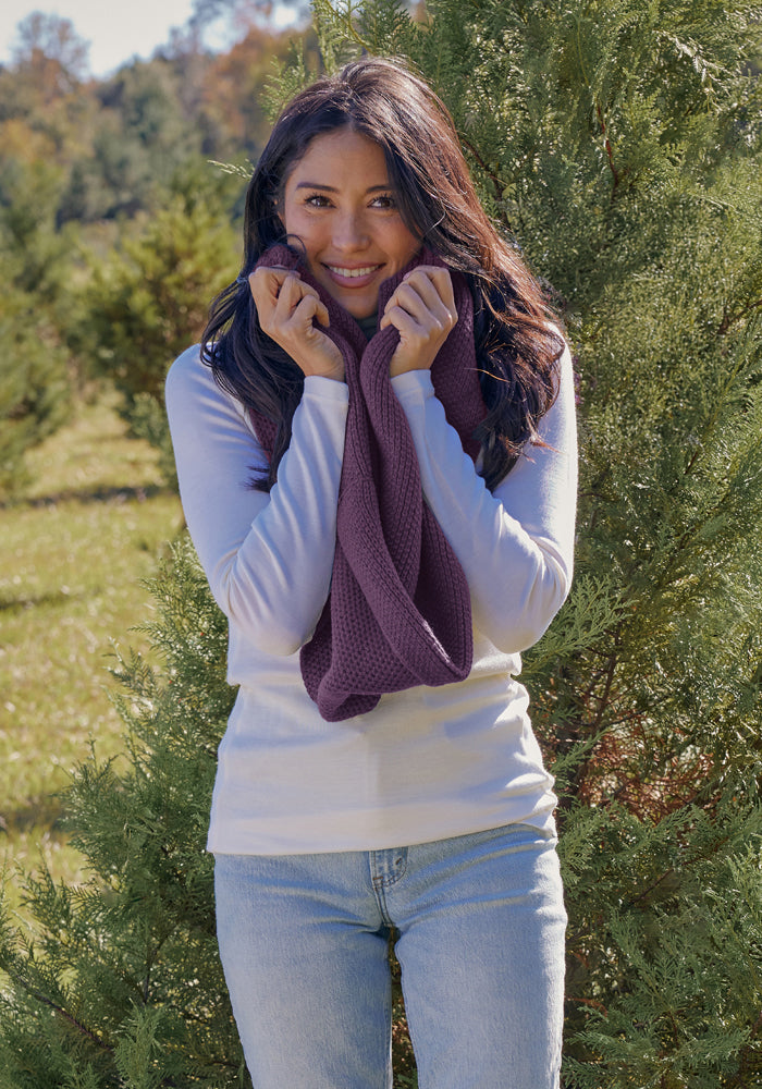 Smiling woman outdoors in a white shirt, holding a Woolx Noelle Infinity Scarf - Purple Iris close to her face.