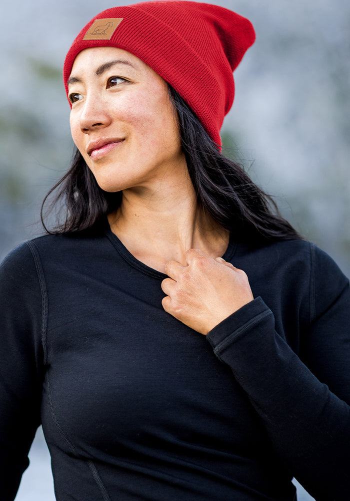 Woman wears Woolx Baylor Beanie in Maraschino Cherry and black shirt outdoors, smiling slightly, hand near collar.