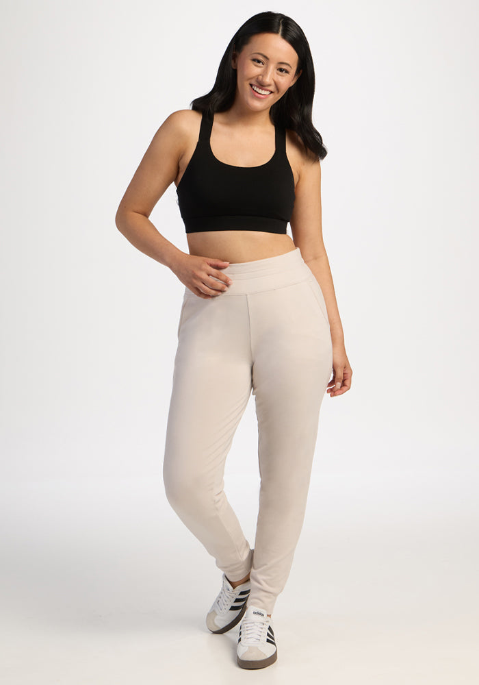 Woman wears Woolx Willa Joggers Petite in Ecru, a black crop top, and white sneakers, smiling against a white backdrop.