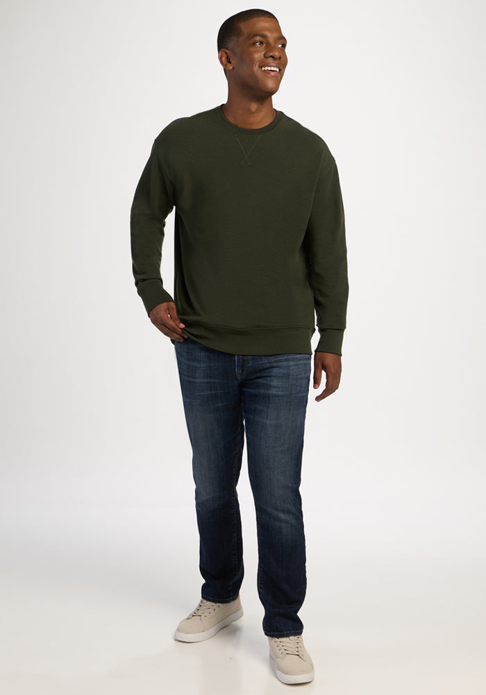 A man in a dark olive green Woolx Miles Crewneck - Duffel Bag, blue jeans, and light-colored sneakers.