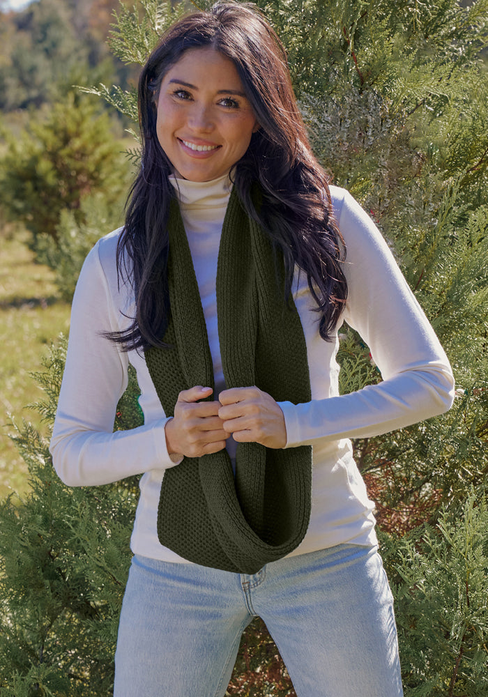 Smiling woman in jeans and white turtleneck outdoors, holding Woolx Noelle Infinity Scarf - Duffel Bag near green foliage.