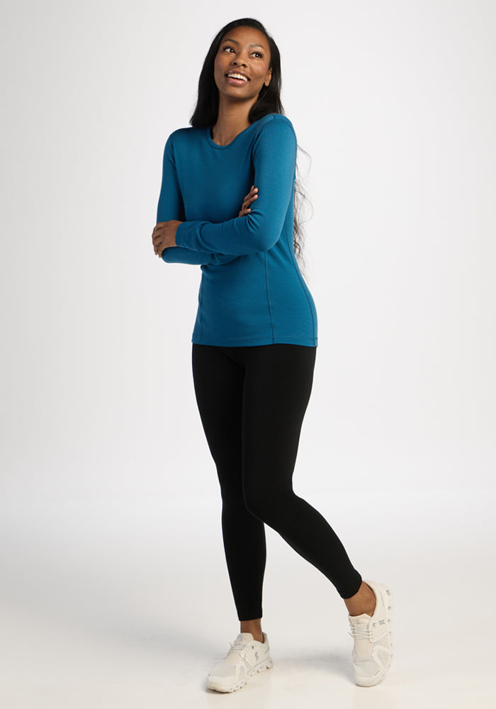 Woman in Woolx Hannah Petite Long Sleeve Top - Blue Pine, black leggings, and white sneakers, smiling against plain backdrop.