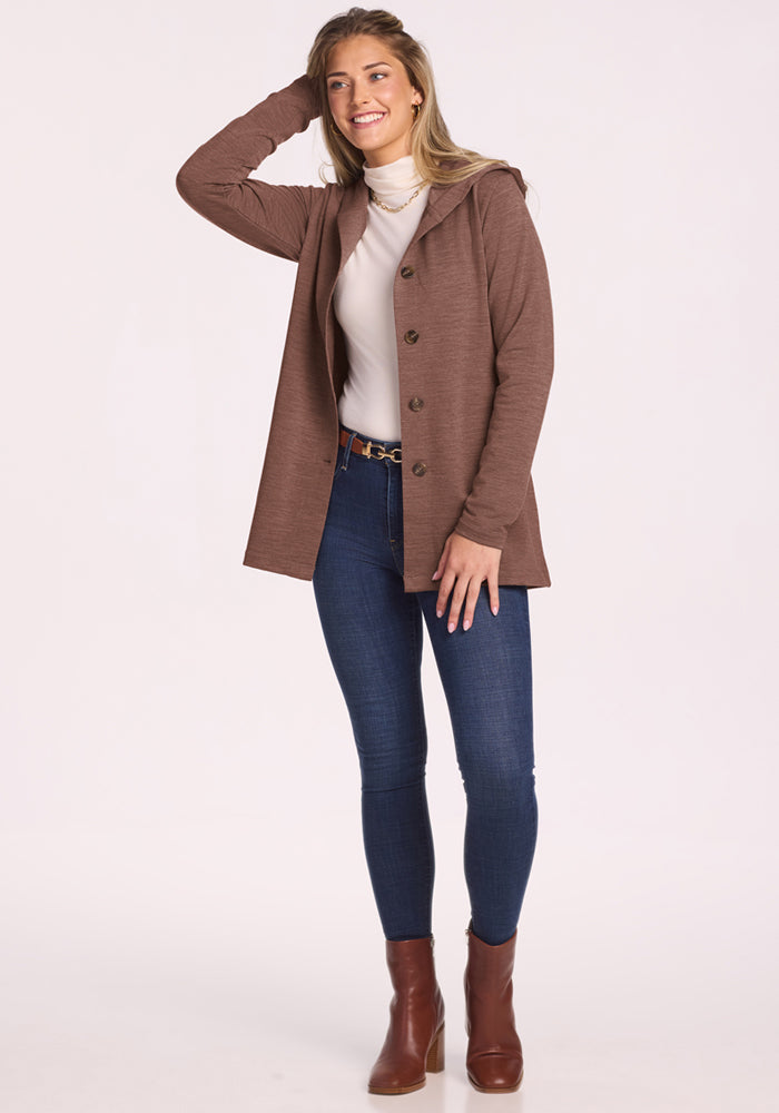 Woman in Edna Hooded Cardigan by Woolx, white turtleneck, jeans, and boots stands smiling with hand on head.