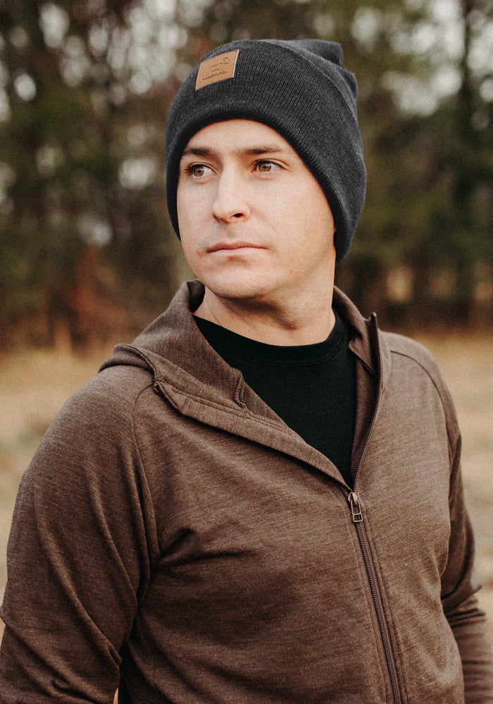 A man wears the Woolx Baylor Beanie in Cinder Melange and a brown zip-up jacket, standing outdoors, looking sideways.