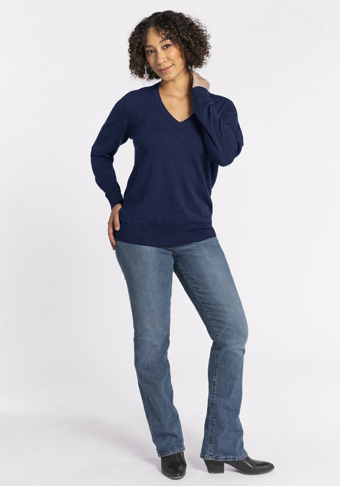 Woman with curly hair wears a Woolx Francesca Sweater in Ocean Cavern, jeans, and black boots against a white background.