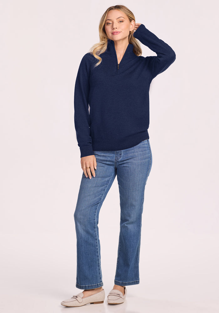 Woman in Woolx Collins Half Zip Sweater - Ocean Cavern, jeans, and loafers stands smiling with a hand on her head.