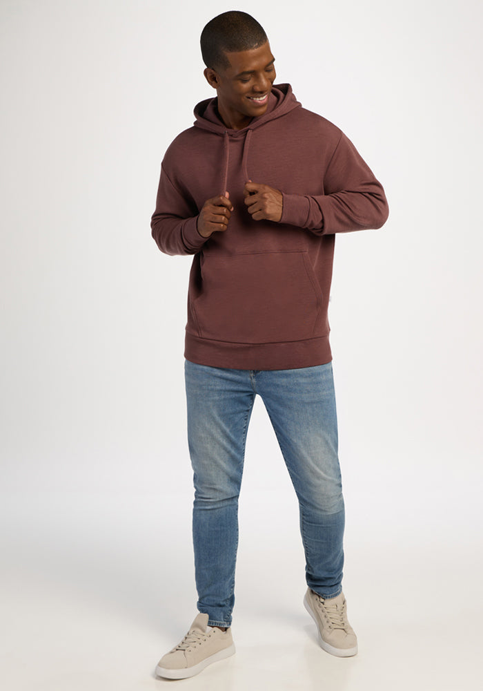 Smiling person wears Woolx Watson Pullover in Canyon Clay, light blue jeans, and beige sneakers against a plain background.