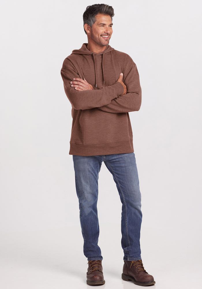 Man in Woolx Chase Hoodie - Sable Heather, blue jeans, and brown boots stands smiling with arms crossed.