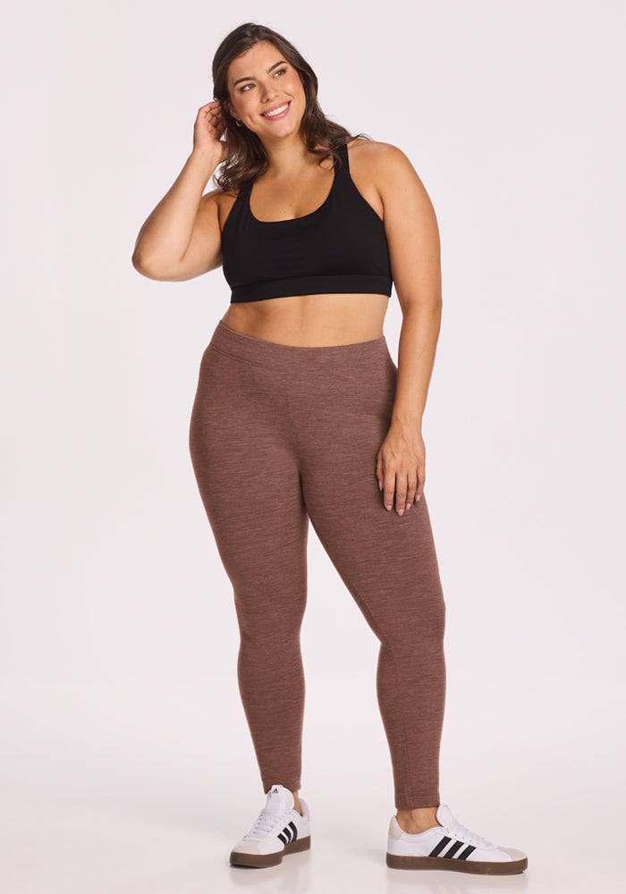 Woman in Woolx Stella Leggings - Sable Heather, black sports bra, and sneakers stands smiling against a white background.