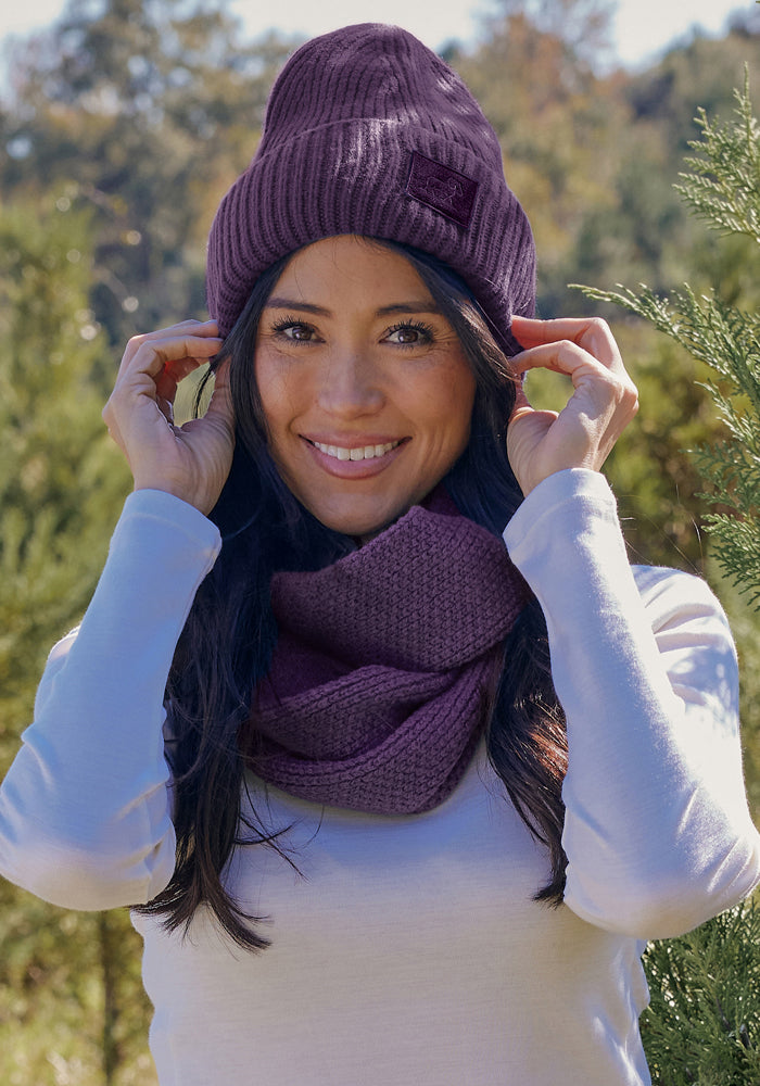 Woman outdoors in the Woolx Ria Chunky Beanie - Purple Iris, smiling and holding her hat with trees behind her.
