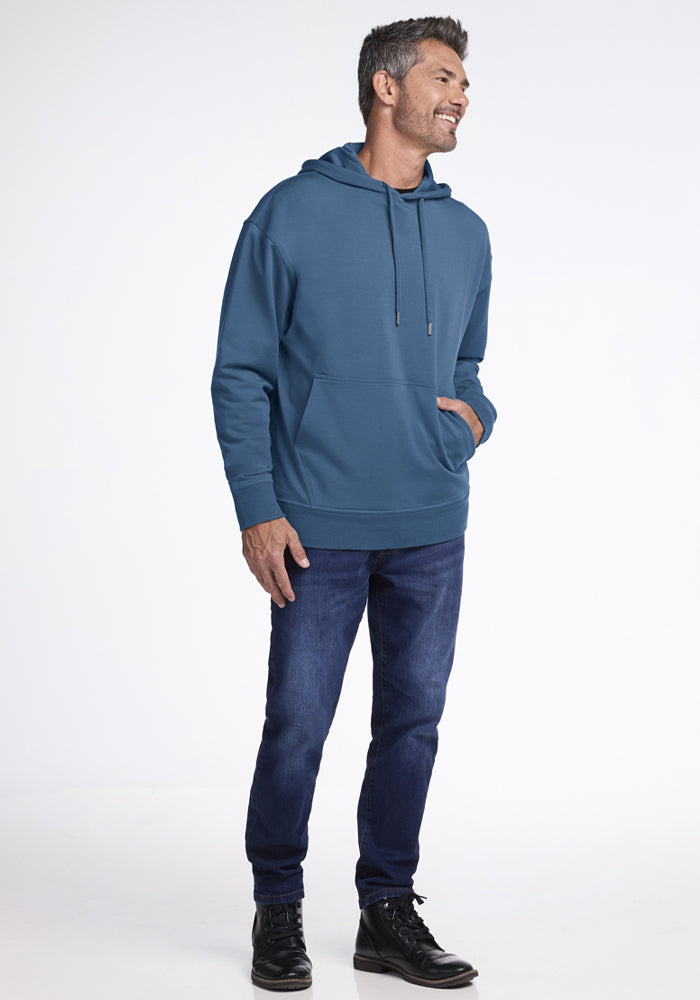 Man in a Woolx Chase Hoodie - Blue Ashes, dark jeans, and black shoes smiles against a white background.
