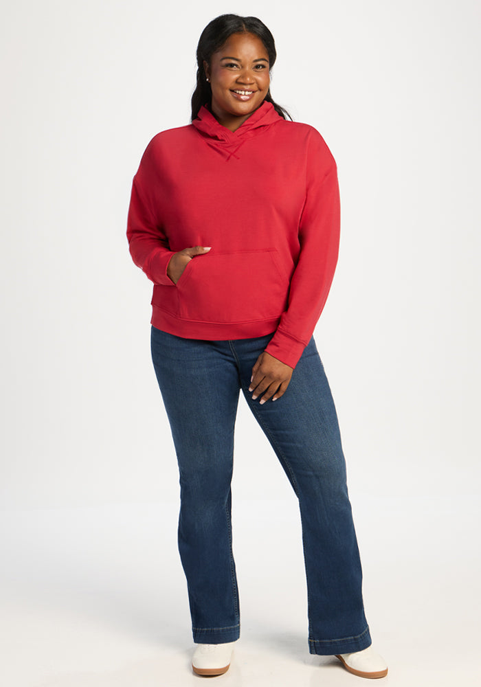 Woman smiles in a Woolx Lizzy Hoodie - Maraschino Cherry and blue jeans, standing against a plain white background.
